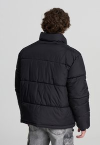 Giacca a vento nera con colletto alto, caratterizzata da un design a trapunta e polsini elastici. Il tessuto è liscio, offrendo un aspetto caldo e isolato.