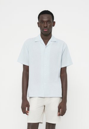 Chemise à manches courtes bleu clair avec col, en tissu texturé et coupe décontractée. Portée avec un short beige.