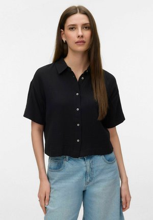 Kobieta z prostymi brązowymi włosami, ubrana w czarną koszulę z krótkim rękawem zapinaną na guziki i jasnoniebieskie dżinsy, stojąca na jednolitym białym tle.