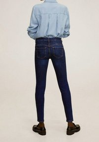 Persona vestida con jeans ajustados de color azul oscuro, camisa de manga larga de color azul claro metida por dentro, y mocasines negros de pie mirando hacia fuera.
