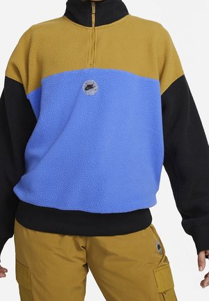 Osoba ubrana w polarowy sweter z blokami kolorów: niebieskim, musztardowym i czarnym oraz pasujące musztardowe spodnie typu cargo z logo Nike.