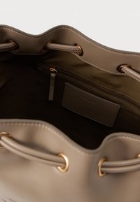 Smėlio spalvos kibirinis krepšys, pagamintas iš lygaus odos, su auksiniais aksesuarais, raišteliu uždaromu ir vidiniais užtrauktuku uždaromu kišenėle, kurioje yra kortelių laikiklis.