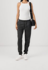 Černé tepláky s elastickými manžetami a nastavitelným šňůrkovým pasem, v kombinaci s bílým topem bez rukávů a bílými teniskami. Doplňkem je malá černá taška.