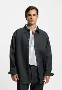 Giovane uomo che indossa una giacca di jeans scura oversize sopra una camicia bianca a righe e jeans scuri, in piedi contro uno sfondo chiaro e uniforme.