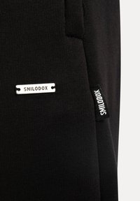 Sorte træningsbukser med ribbet tekstur. Har sølvfarvet "SMILODOX" branding og et stofmærke med samme navn på siden.