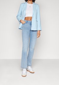 Světlomodrý blejzr, bílý crop top, světlomodré zvonové džíny a bílé tenisky s béžovými akcenty, vše vystaveno na neutrálním pozadí.