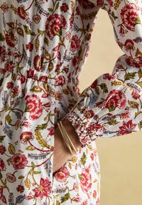 Blumenkleid aus weißem Stoff mit roten und blauen Mustern. Die elastischen Ärmel und der Taillenbund bieten Struktur und Komfort.