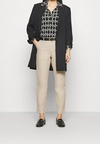 Cappotto nero su una blusa con motivo a link bianchi, pantaloni beige attillati e mocassini neri. Presenta una cintura nera con una fibbia circolare.