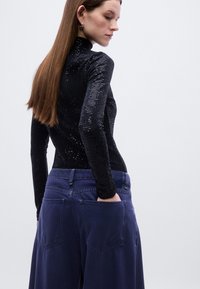 Μαύρο τοπ μακρυμάνικο με λαμπερή υφή, σε συνδυασμό με χαλαρά σκούρα μπλε παντελόνια με πίσω τσέπες και ραφές.