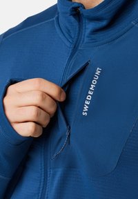 Niebieska kurtka zapinana na zamek z wysokim kołnierzem, charakteryzująca się eleganckim stylem i widocznym logo. Materiał wydaje się gładki, a rękawy mają otwory na kciuki.
