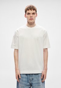 Tricou alb cu mâneci scurte din bumbac, cu gât rotund și un mic logo pe partea stângă a pieptului, asortat cu blugi bleu deschis.