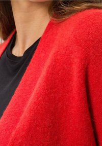 Czerwona, miękka kardigan z dzianiny z dekoltem w serek, o puszystej fakturze i otwartym przodzie. Nałożona na czarną koszulę.