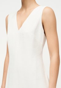 Αμάνικο λευκό φόρεμα με διακριτή υφή, με λαιμόκοψη V, που φοριέται από άτομο με γυμνά χέρια και λαιμό ορατό.