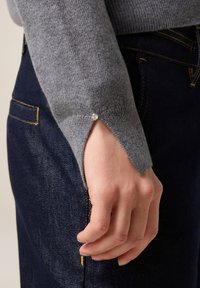 Szary sweter z dzianiny z delikatnym otwarciem przy mankiecie. Manicet ma niewielki akcent, a sweter zestawiony jest z ciemnymi dżinsami.