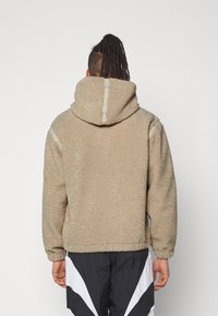 Beige fleecejacka med huva, som har en mjuk yta, elastiska mudd och sömdetaljer längs ryggen och axlarna.