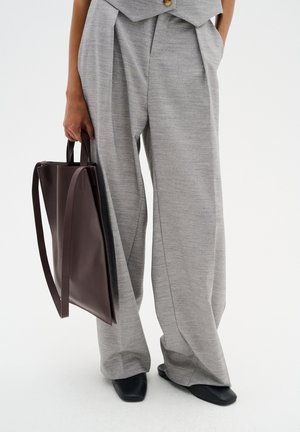 Grau gefaltete Hosen aus strukturiertem Stoff, kombiniert mit einer dunkelbraunen Ledertasche mit kurzen und langen Griffen. Schwarze flache Schuhe.