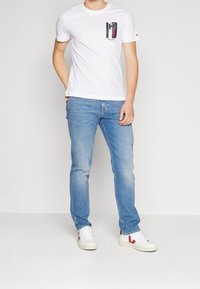 Bílé tričko s krátkým rukávem a grafickým logem na hrudi, kombinované se světle modrými džínami a bílými teniskami s červenými detaily.