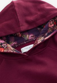Felpa con cappuccio rosso scuro che mostra l'interno del cappuccio foderato con tessuto floreale e l'etichetta visibile del marchio Ted Baker London.