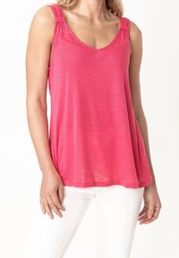 Top leggero rosa senza maniche con scollatura a V, tessuto texturizzato e dettagli arricciati sulle spalline, abbinato a pantaloni bianchi.