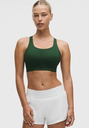 Femme en forme debout, portant un soutien-gorge de sport vert foncé et un short de sport blanc, sur un fond clair uni.