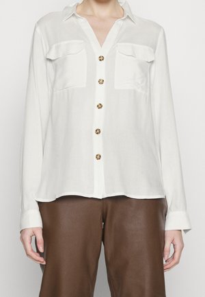 Chemise blanche à manches longues boutonnée avec deux poches poitrine portée avec un pantalon en cuir marron.