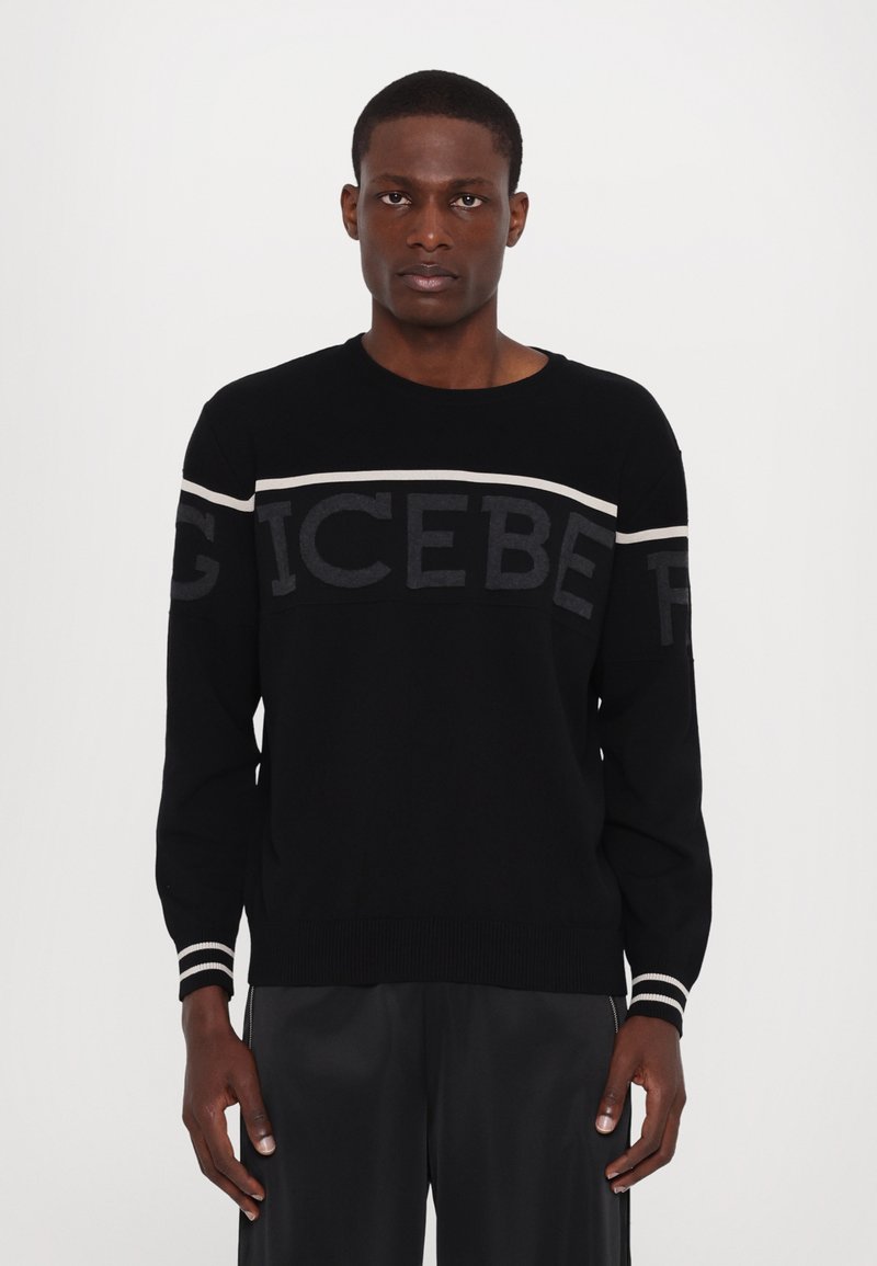 Pull noir avec des lettres grises claires et foncées sur la poitrine. Encolure et manches côtelées avec des bandes blanches. Matière en tricot douce.
