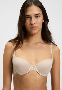 Vaaleanbeige pehmustettu rintaliivi, jossa on alumiinituki, sileä pinta, säädettävät hihnat ja pitsikoristelu keskiosassa.