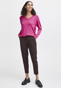 Rosa v-ringad långärmad tröja tillsammans med bruna avsmalnande byxor och svarta loafers. Tyget ser lätt och med en slät yta.