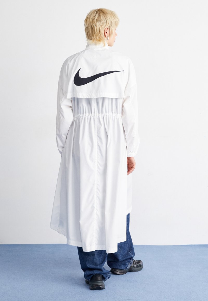 Persoon met kort blond haar, gekleed in een lange witte Nike-jack, blauwe spijkerbroek en zwarte sneakers, staande op een blauwe vloer tegen een witte muur.