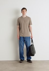 Brun bomulls t-skjorte med en liten logo, kombinert med blå denimjeans. Modellen bærer en svart totemappe og har på grå sko.