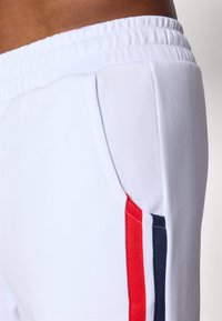 Vita joggingbyxor med elastisk midja. Har sidofickor och röda samt marina ränder längs ytterkanterna. Mjuk och slät textur.