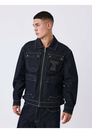 Jeune homme portant une veste en jean foncée avec le texte "CHANGE THE GAME" sur la poche poitrine, regardant vers sa gauche sur un fond uni.