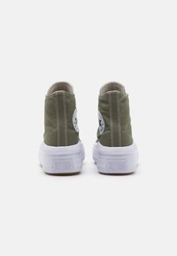 Green canvas high-top sneakers with white rubber soles, featuring a circular logo patch on each side and contrasting stitching details.