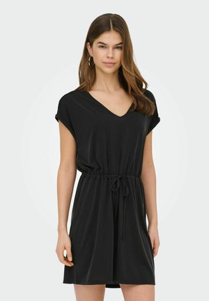 Noor naine, kes kannab musta lühikeste varrukatega V-kaelusega kleiti, millel on nöörvöö vöökoht, seisab ühevärvilise heleda tausta ees.