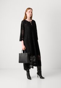 Black lace dress with long sleeves, cutout patterns, and a tie neckline, paired with a textured black handbag and slim heeled boots.