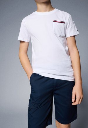 Niño con una camiseta blanca con un bolsillo a rayas y pantalones cortos azul oscuro, una mano en el bolsillo, de pie frente a un fondo gris liso.