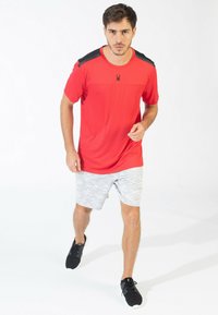 Camiseta atlética roja con acentos negros en los hombros, combinada con pantalones cortos de camuflaje gris claro y zapatillas negras. El modelo está en una postura de caminata.
