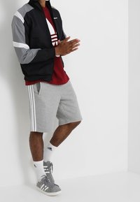 Man wearing black and gray jacket, red Adidas shirt, gray Adidas shorts, white Adidas socks, and gray Adidas sneakers standing against white wall.