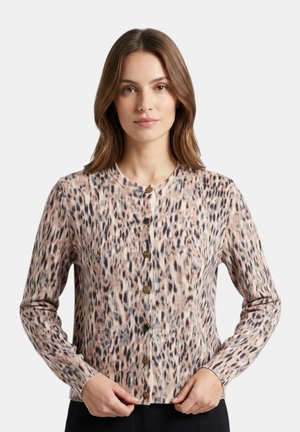 Donna con capelli castani fino alle spalle indossa un cardigan beige e nero con stampa leopardata e bottoni, in piedi davanti a uno sfondo chiaro e uniforme.