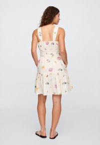 Vestido de verão com padrão floral em tecido bege claro, com alças largas, corpete franzido e saia em camadas. Costas abertas e comprimento até ao tornozelo na modelo.