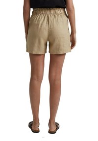 Vue arrière d'une personne portant un short beige à taille élastique, un haut noir et des sandales noires, debout devant un fond blanc.