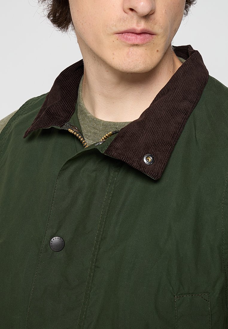 Mann trägt eine dunkelgrüne Jacke mit braunem Cordkragen und einem olivgrünen Hemd darunter, leicht nach rechts gewandt, mit geschlossenen Lippen.