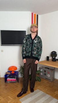 Rakstains melns kardigans ar zaļām un ziedu akcentiem, apvienots ar brūnām platajām biksēm un tumšām kurpēm. Redzami neitrāli fona elementi.