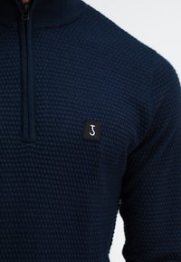 Donkerblauwe gebreide trui met een gestructureerd patroon, kwart ritssluiting en een klein zwart logo-embleem met een gestileerde "J" op de borst.