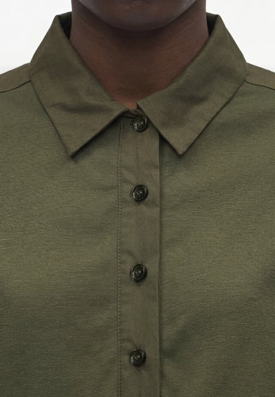 Κοντινό πλάνο ενός σκούρου δέρματος ατόμου που φοράει ένα ελιάς πράσινο πουκάμισο με γιακά, κουμπωμένο με τέσσερα ορατά σκοτεινά κουμπιά.