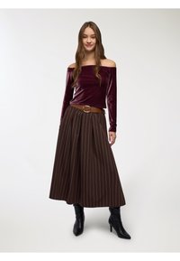 Top in velluto bordeaux con scollo off-the-shoulder abbinato a una gonna midi plissettata a righe marroni, accentuata da una cintura marrone con fibbia rotonda e stivali neri con tacco.