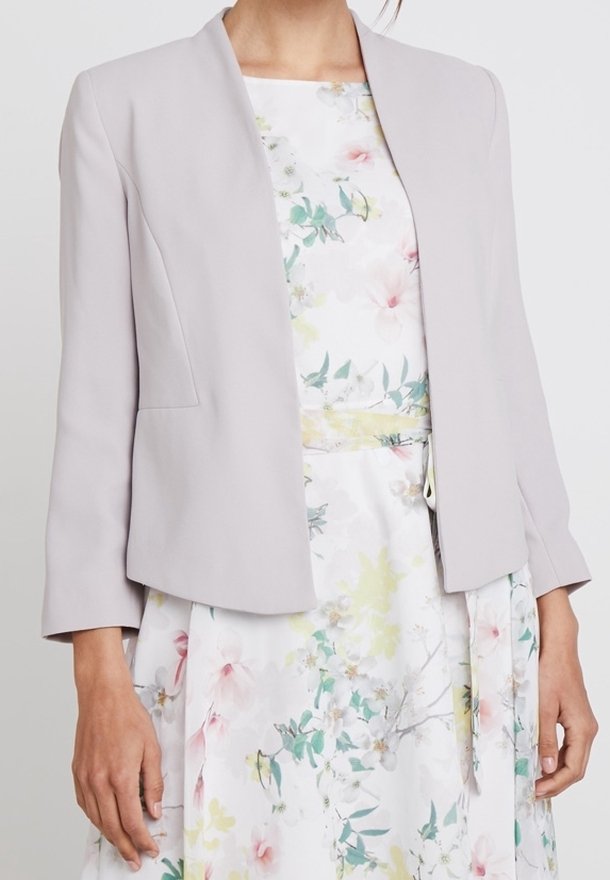 Veste grise claire ajustée avec une finition lisse, dotée d'un col rond et de manches longues, associée à une robe à imprimé floral.