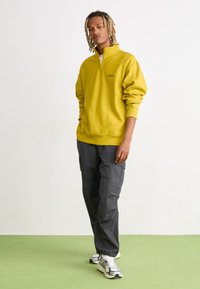 Geel rits-trui met hoge kraag, gecombineerd met donkergrijze cargobroek. Model draagt witte en zwarte sneakers. Eenvoudig ontwerp, relaxte fit.