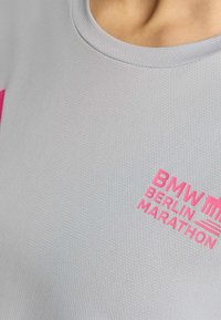 Camisa deportiva gris de manga corta y cuello redondo. Presenta el texto en rosa "BMW BERLIN MARATHON" y el logo, fabricada con un material ligero y texturizado.