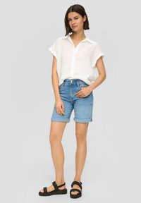 Biała, krótka koszula na guziki z krótkim rękawem, jasnoniebieskie, podwinięte szorty dżinsowe, czarne sandały na paski, modelka stojąca na szarym tle.